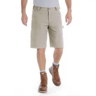 Mężczyzna nosi beżowe spodenki Carhartt Rigby Dungaree Short, stojąc na białym tle. Ma na sobie również szare skarpetki i brązowe skórzane buty.