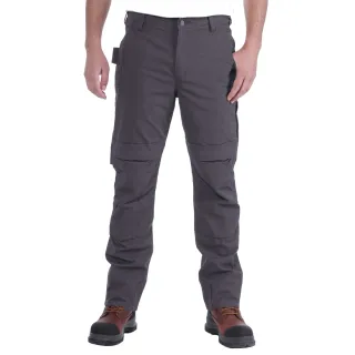 Mężczyzna nosi szare spodnie robocze Carhartt Full Swing Steel Multi Pocket Pant z wieloma kieszeniami. Na nogach ma brązowe buty robocze z czarnymi noskami.